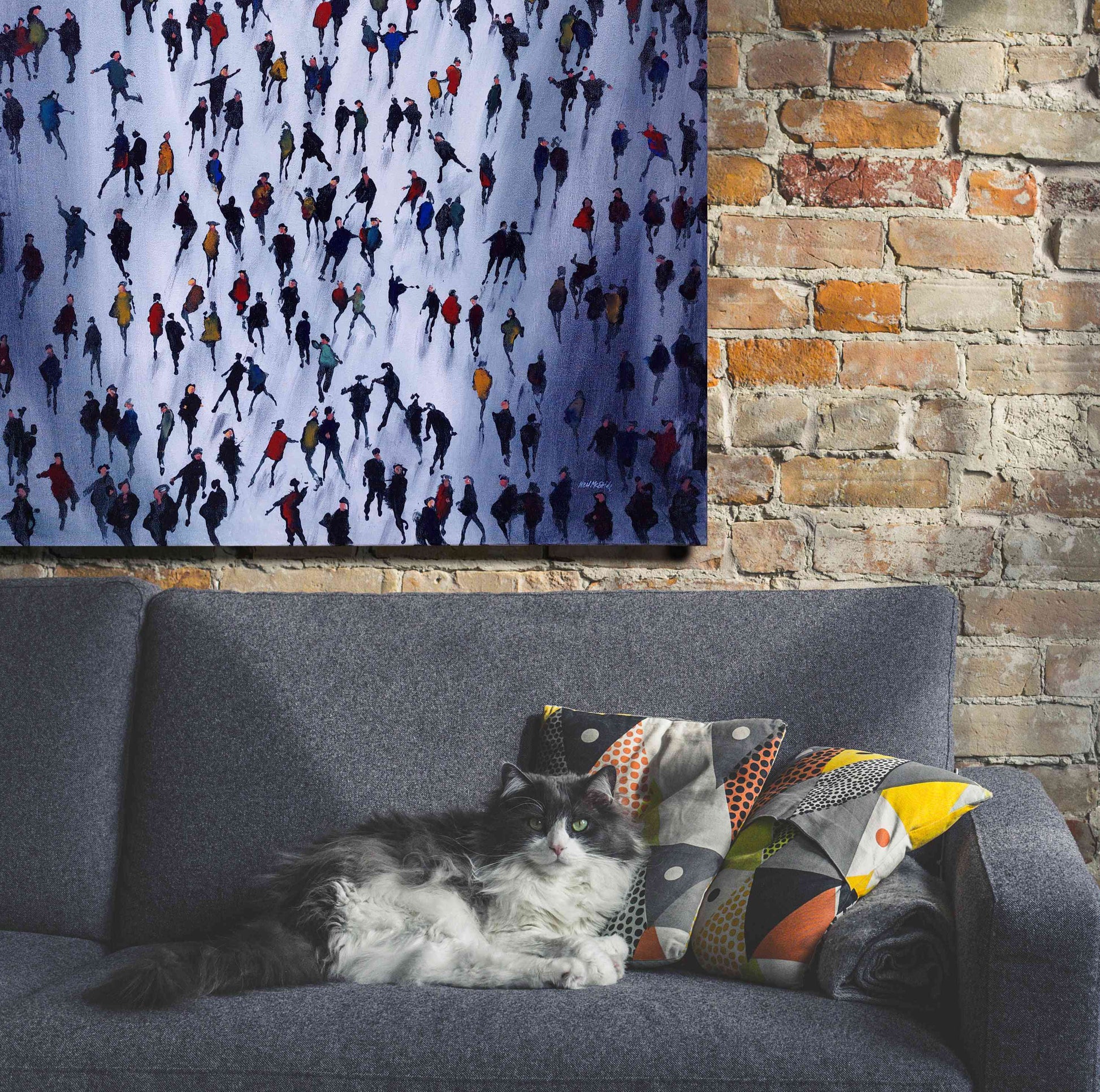 Cat lying on a grey sofa with colourful  patterned cushions. A large crowd painting by Neil McBride is displayed on a bare brick wall behind the sofa.